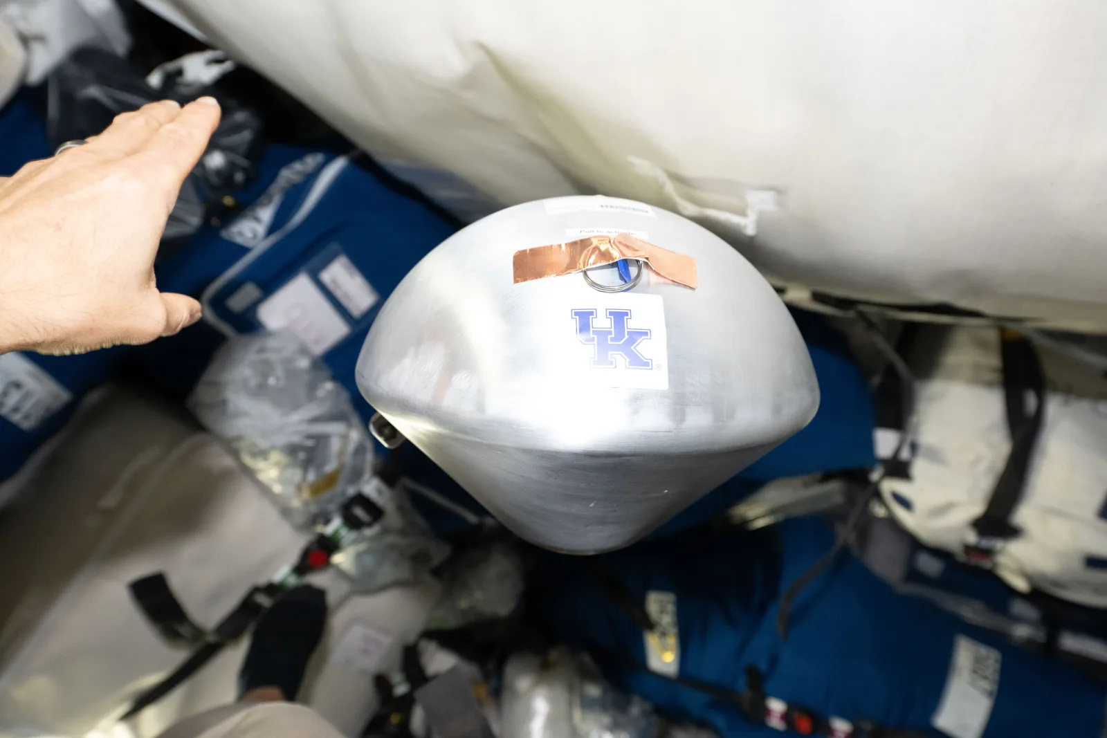 A metallic research capsule floating inside the cargo compartment, with a hand at upper left and stowed equipment behind.